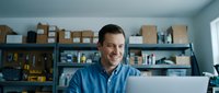 Middle-aged Caucasian businessman smiling while working on laptop in modern warehouse office with shelves and cardboard boxes in background.