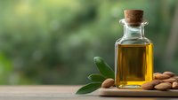Golden almond oil in glass bottle with cork stopper and fresh almonds on wooden table against blurred green natural background for skincare and cooking.