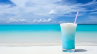 Tropical blue cocktail drink with pink straw on white sand beach against turquoise ocean water and cloudy sky for summer vacation and resort concepts.
