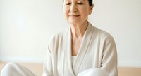 Mature Asian woman in white robe sitting peacefully with eyes closed, expressing wellness, mindfulness and relaxation for spa, healthcare and lifestyle content.