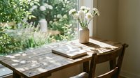 Rustic wooden table with white flowers in a vase and an open book by a sunlit window overlooking a lush green garden.