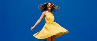 Young woman in flowing yellow dress dancing against bright blue sky background with hair in motion expressing joy and freedom.