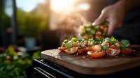 Fresh caprese salad preparation with hands sprinkling herbs over sliced tomatoes and mozzarella on wooden cutting board, backlit by golden sunlight in outdoor setting.