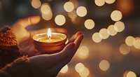 Female hand holding burning candle in clay pot against blurred golden bokeh lights background creating spiritual peaceful atmosphere for meditation and prayer.