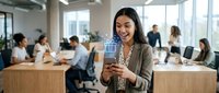 Young woman with dark hair and South Asian ethnicity, wearing a beige blazer and patterned blouse, smiling while holding a digital tablet in a modern office environment with colleagues working at wooden desks.