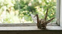 Paper origami crane sitting on wooden windowsill with soft natural light and blurred green foliage background creating peaceful zen atmosphere.