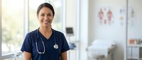 Smiling Hispanic female nurse in navy blue scrubs with stethoscope standing in modern hospital clinic ready for patient care and medical assistance.