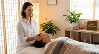 Professional female therapist performing reiki healing treatment on relaxed client in serene wellness spa room with plants and natural lighting.