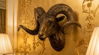 Mounted goat head with curved horns on golden damask wallpaper wall, flanked by white pleated lamps, vintage taxidermy decor for luxury interior design and home styling.