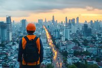 Young Asian male traveler with orange hardhat and backpack overlooking modern city skyline at sunset, illuminated street stretches to horizon amid skyscrapers and urban landscape.