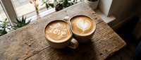 Two ceramic mugs with latte art on a rustic wooden table, one featuring the word Love and the other a heart, cozy cafe atmosphere.