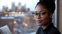 Professional African American businesswoman with glasses working on laptop in modern office with city skyline background during evening hours.