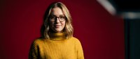 Young blonde Caucasian woman in yellow turtleneck sweater and glasses smiling warmly in modern office setting with dramatic lighting for business portraits.