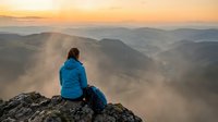 Woman with reddish-brown hair wearing a blue jacket sits on a rocky mountain peak at sunrise, gazing over misty valleys and distant hills during golden hour.