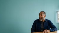 Middle-aged man with dark skin, gray hair, and beard wearing navy blue shirt against turquoise wall. Suitable for business, corporate, professional portraits, and editorial use.