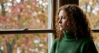 Young woman with curly hair in green sweater looking thoughtfully out window during autumn season with colorful foliage background for lifestyle content.