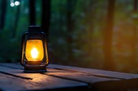 Glowing vintage oil lamp on wooden surface at dusk, warm light illuminating dark forest background creates atmospheric outdoor scene.