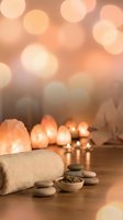 Serene spa setting with salt lamps, stacked stones, rolled towel, and herbs on wooden surface, warm bokeh lights in background.