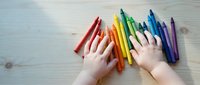 Child hands reaching for colorful crayons arranged in rainbow pattern on wooden table surface for creative art projects and educational activities.