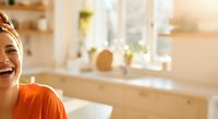 Young woman with South Asian ethnicity wearing orange shirt and headband, smiling in modern bathroom with natural lighting and minimalist decor, suitable for wellness and lifestyle content.