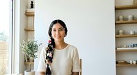 Young woman with long dark hair in braid, olive skin tone, wearing casual cream t-shirt, sitting indoors by window with shelving and plants, portrait style for lifestyle and wellness content.