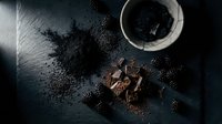 Dark chocolate pieces with cocoa powder scattered on black slate surface creating moody culinary composition for gourmet cooking and baking projects.