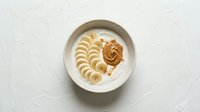 Overhead view of creamy yogurt bowl with sliced banana and peanut butter swirl on white textured surface, healthy breakfast or snack option.