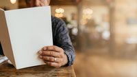 Mature man with dark skin holding blank white card at wooden table in blurred indoor setting, suitable for voting, surveys, feedback, or promotional messaging campaigns.