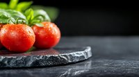 Fresh ripe tomatoes with water drops and green leaves on black stone board against dark background, macro food photography with copy space.