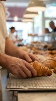 Male baker with medium skin tone arranging freshly baked croissants on cooling rack in professional bakery kitchen with blurred staff and equipment in background.