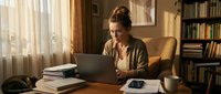 Professional woman working on laptop at wooden desk in cozy home office with books and warm natural lighting for remote work and productivity concepts.