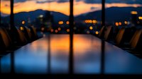Empty modern conference room with reflective table at sunset, city lights and mountain silhouettes visible through panoramic windows, creating dramatic evening atmosphere.