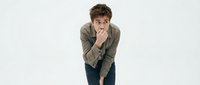 Young man with brown hair and light skin tone wearing casual khaki jacket and dark jeans, displaying anxious expression with hand near mouth against white background.