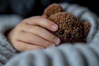 Child hand gently holding brown teddy bear in cozy bed setting. Comfort, care, childhood memories, bedtime routine, soft toy companionship concept.