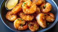 Crispy breaded shrimp garnished with fresh herbs and sesame seeds served on dark plate with dipping sauce, close up view of appetizing seafood dish.