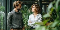 Caucasian couple smiling at each other in modern office space with green plants, representing workplace relationships and positive corporate culture.