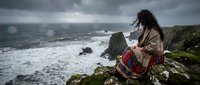 Young woman with long dark hair in traditional ethnic clothing sits on dramatic coastal cliff overlooking stormy ocean waves under moody gray sky.