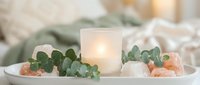 Cozy spa arrangement with glowing candle, eucalyptus leaves and pink salt crystals on white tray creating peaceful wellness atmosphere for relaxation.