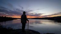 Elderly man with walking stick silhouetted against dramatic sunset sky reflected in calm lake water. Peaceful retirement lifestyle and nature contemplation.