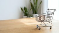 Miniature shopping cart on wooden table with potted plant in background, representing retail commerce and consumer spending for business concepts.