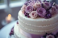 Elegant wedding cake decorated with pink and purple roses, pearl details, and berries on white fondant, captured in soft romantic lighting with blurred background.