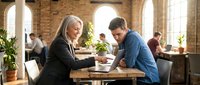 Mature woman with long gray hair and glasses wearing black blazer shaking hands with young man in blue shirt at wooden table in modern brick office space with colleagues working in background.