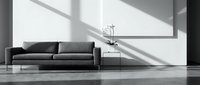 Dark gray upholstered sofa with metal legs positioned in minimalist interior space with white walls, floor lamp, and natural light casting geometric shadows.