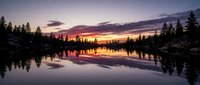 Serene lake at sunset with vibrant orange and purple sky reflecting on calm water, surrounded by evergreen forest and rocky shores, ideal for nature and landscape backgrounds.
