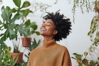 Young African American woman with natural curly hair smiling joyfully among houseplants, wearing casual brown turtleneck sweater against white wall background.