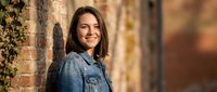 Young Caucasian woman in denim jacket smiling confidently against rustic brick wall with autumn ivy, warm natural light, casual outdoor portrait.