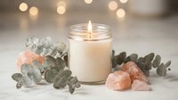 Lit candle in glass jar surrounded by eucalyptus leaves and pink salt crystals creating peaceful spa atmosphere for relaxation and wellness.