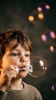 Young boy with brown hair blowing soap bubbles against dark background with colorful bokeh lights creating magical childhood moment for family content.