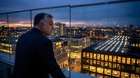 Middle-aged man with gray hair and olive complexion, wearing dark business attire, gazing out from high-rise window at illuminated cityscape during twilight hour for corporate leadership and urban lifestyle concepts.