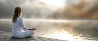 Young woman practicing meditation in lotus position on wooden dock by serene lake at sunrise with misty mountains creating peaceful wellness atmosphere.